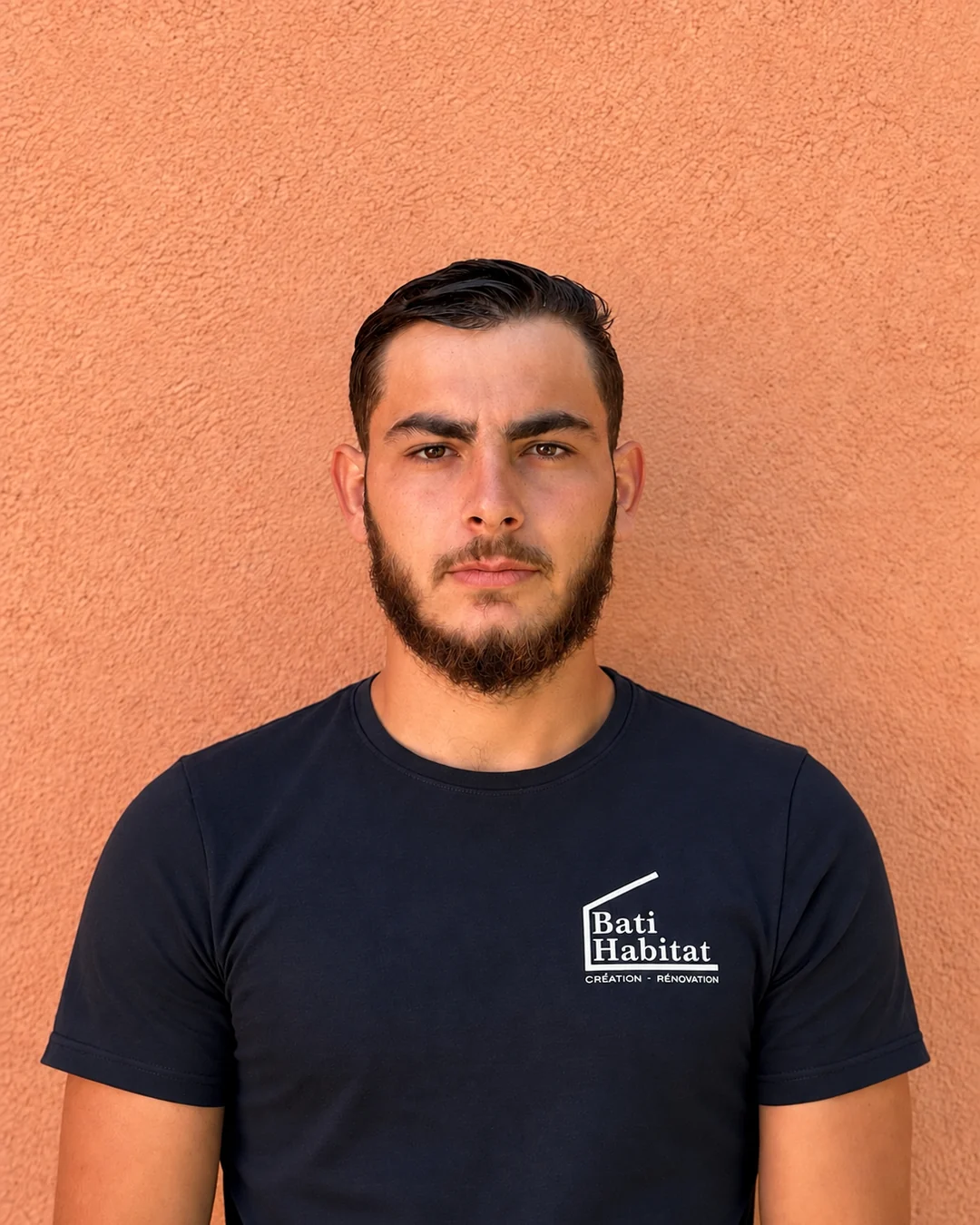 Portrait de Kilian Pasqual, artisan chez Bati Habitat, en t-shirt noir floqué du logo de l'entreprise devant un mur orangé